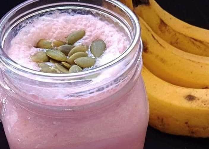 A glass of pink banana rose smoothie topped with seeds next to fresh bananas.