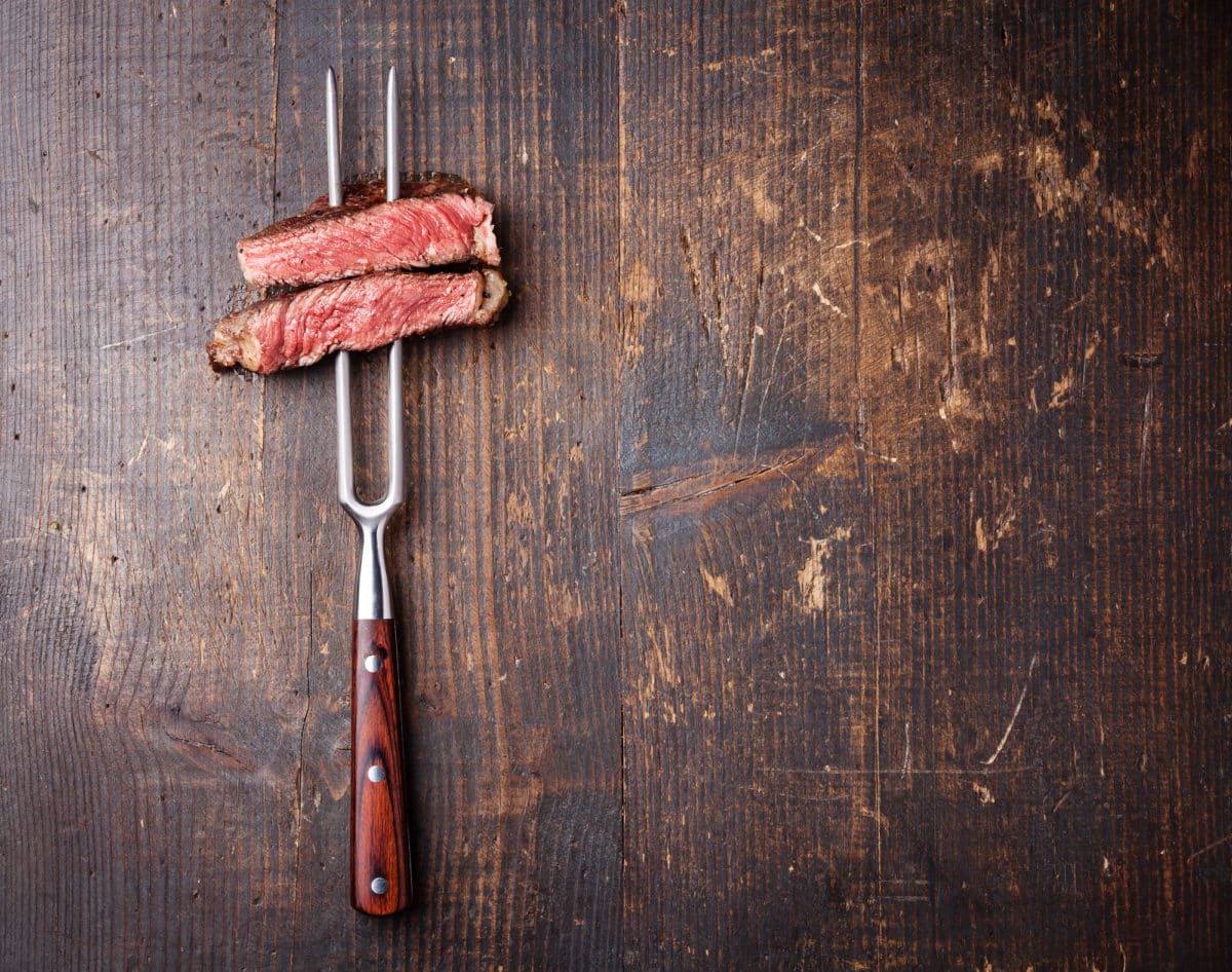 Slices of beef steak on a metal fork against a wooden backdrop.