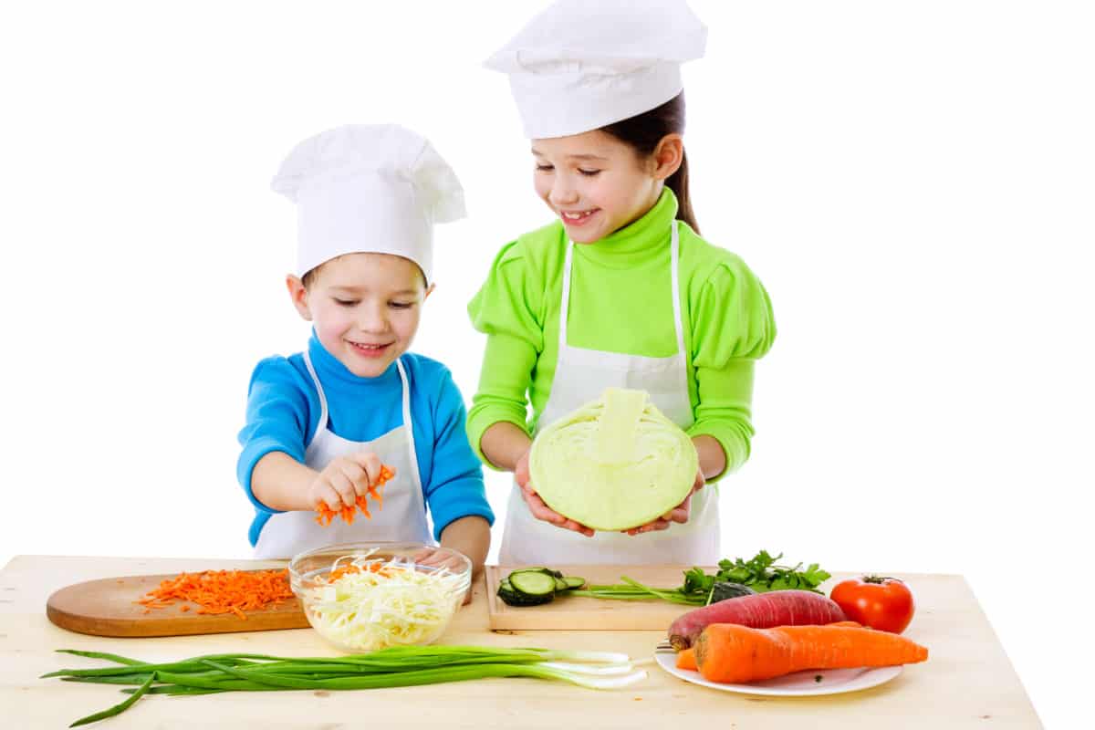 Two children in chef hats smiling as they prepare a fresh salad.