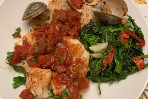A plated dish of Cod Puttanesca with clams, broccoli rabe, and brown rice.