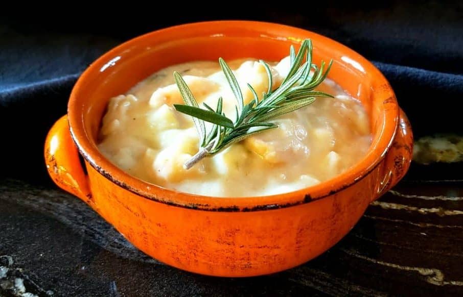 A creamy seafood chowder with chunks of seafood and a sprig of rosemary in an orange bowl.