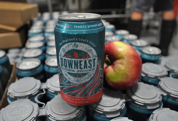 A Downeast Cider House can next to a fresh apple on a stack of Downeast cider cans.