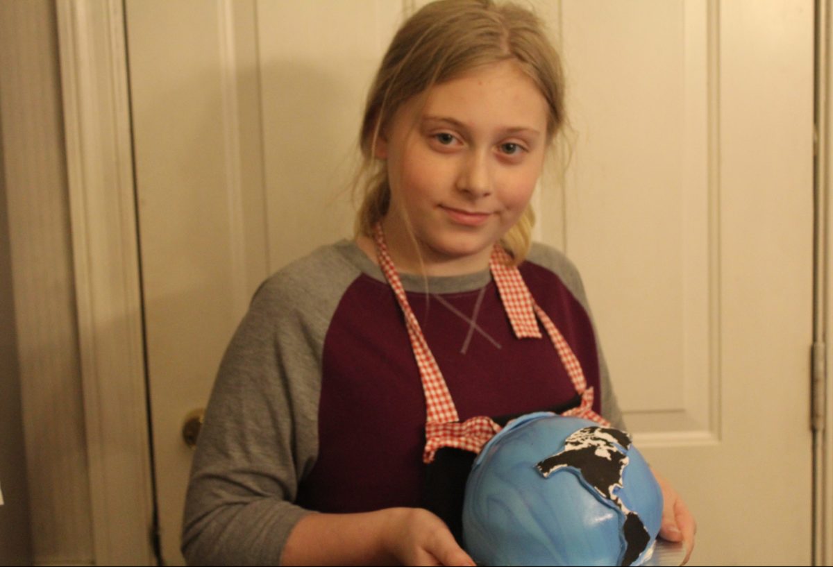 A young girl proudly holding a globe-themed cake in a kitchen setting.