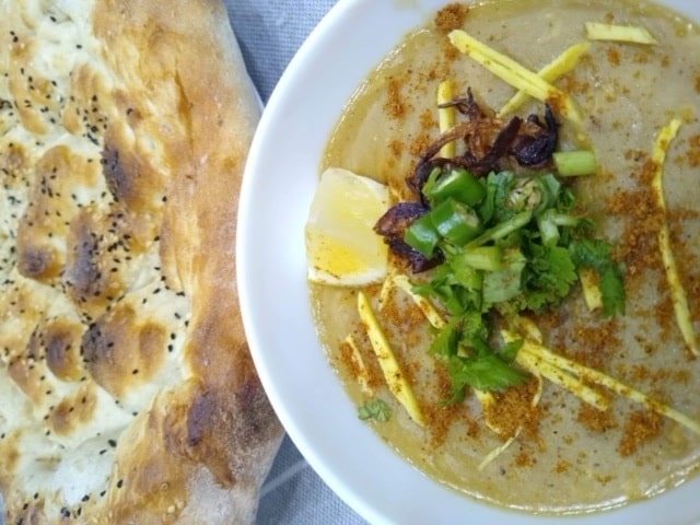 A bowl of haleem garnished with green herbs, fried onions, and lemon slices.