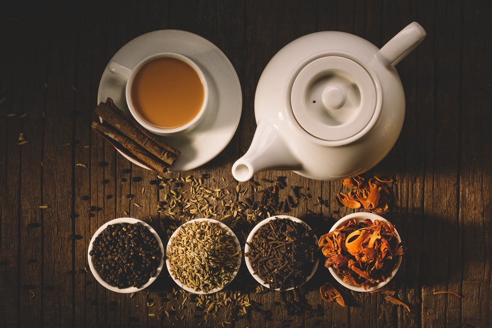 White teapot with cups and assorted tea leaves on a wooden table.