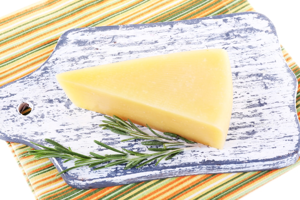 Block of parmesan cheese next to a sprig of rosemary on a rustic wooden board.