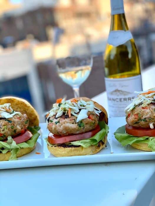 A row of tuna burgers on buns, with lettuce and tomato and topped with coleslaw.