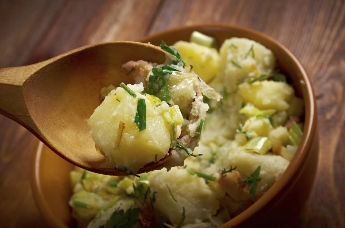 A wooden spoon serving German potato salad with greens and onions.