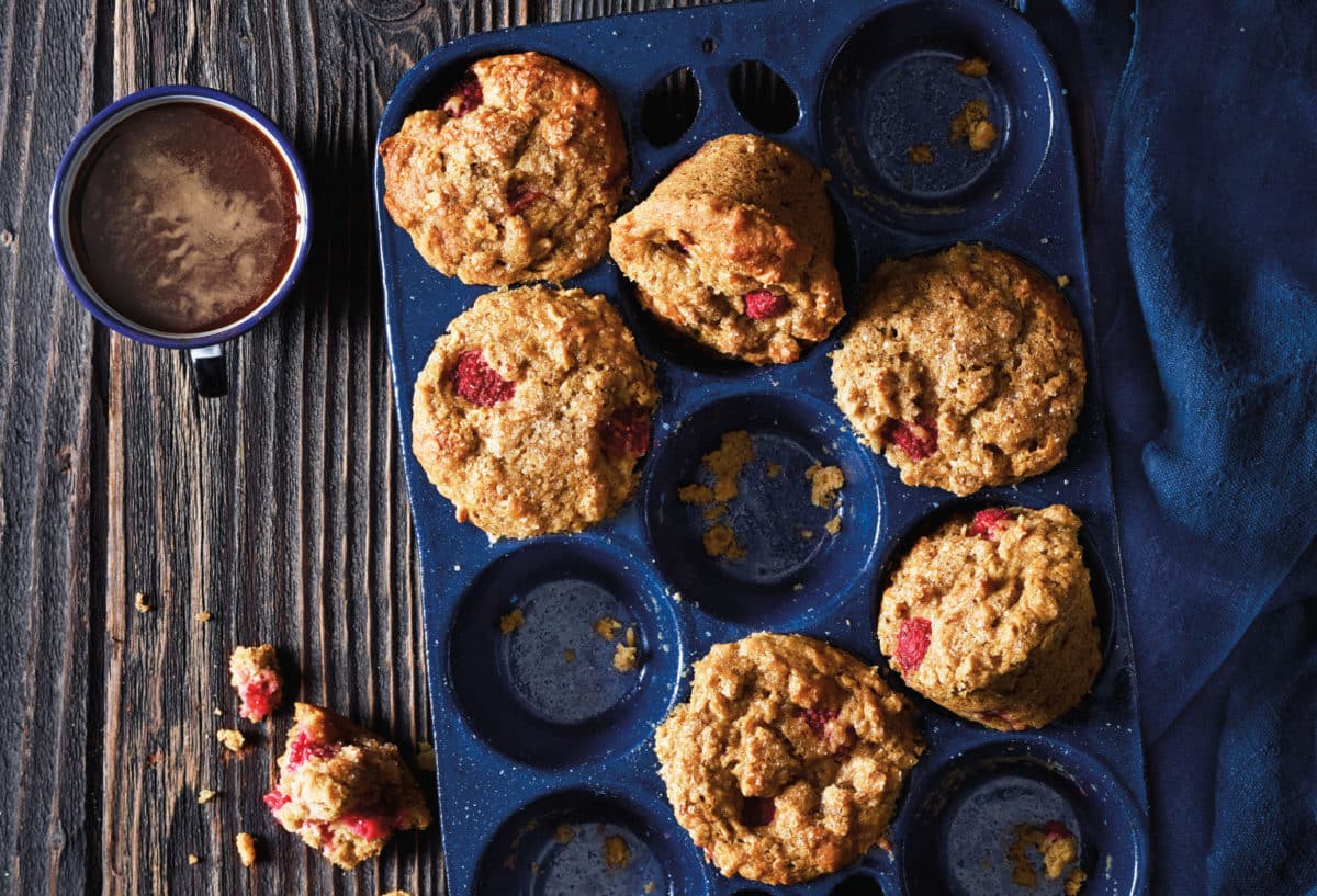 A blue muffin pan filled with golden-brown Raspberry Oatmeal Muffins, with one muffin tilted on its side, next to a steaming cup of cocoa.