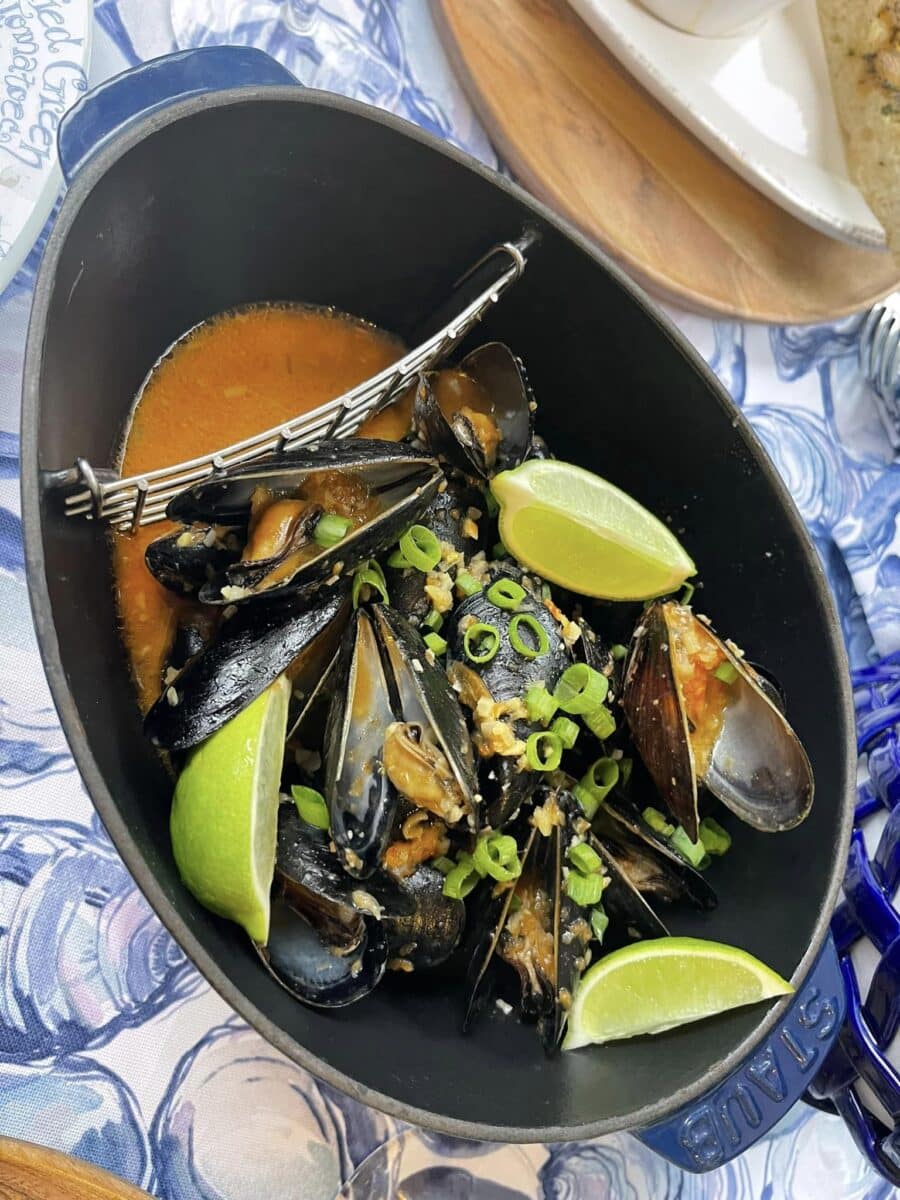 Mussels in the shell in a red curry broth with sliced scallions, herbs, and lime wedges.