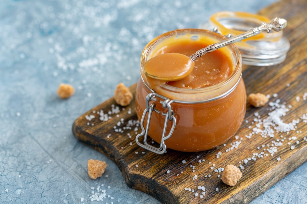 Glass jar filled with caramel sauce, a spoon inside, and scattered salt and brown sugar around the jar.