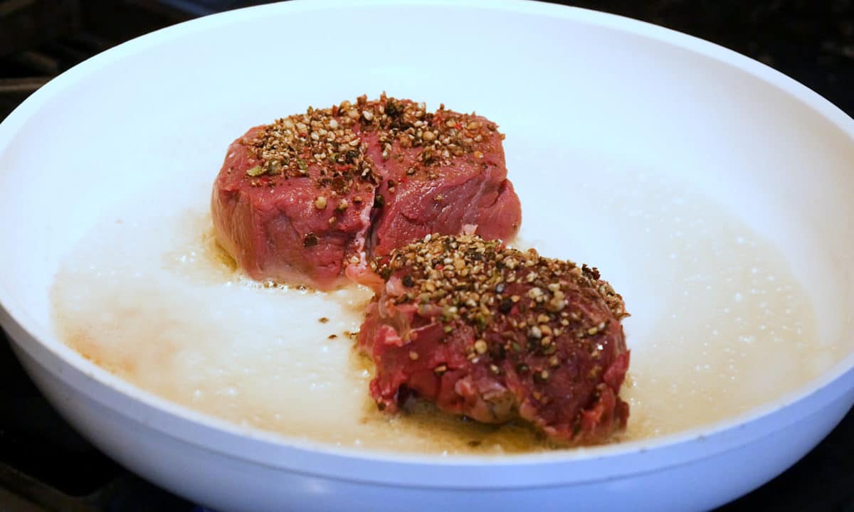 Raw buffalo tenderloin sprinkled with crushed peppercorns, ready for searing in a buttery pan.