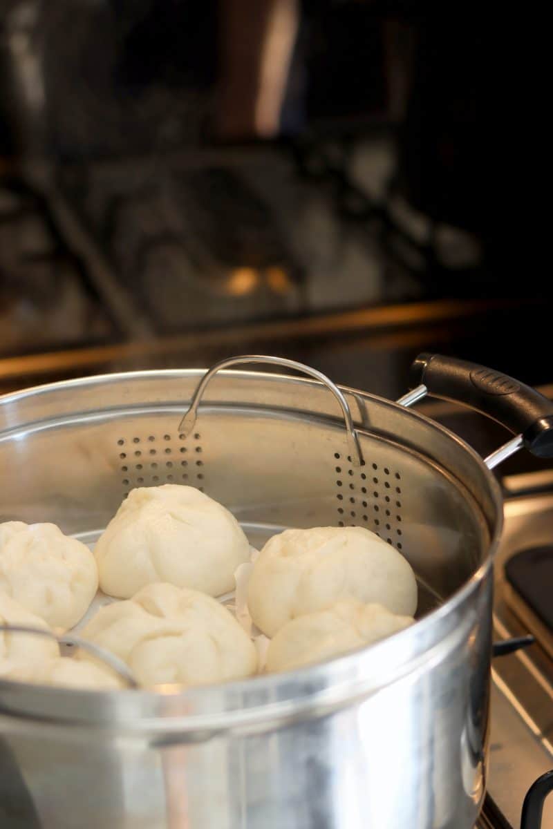 White buns being steamed in a stainless steel steamer on a stove.