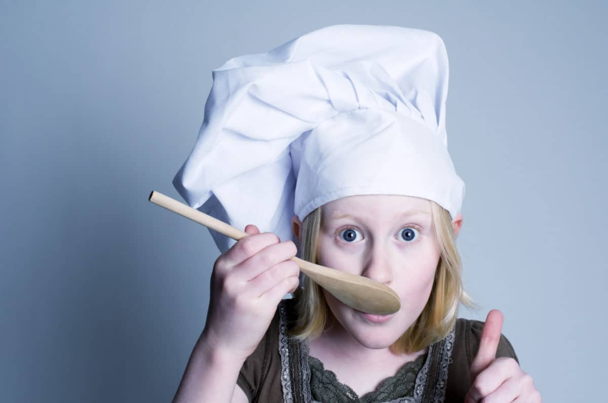 Young chef tasting food with a wooden spoon, giving a thumbs-up in approval.