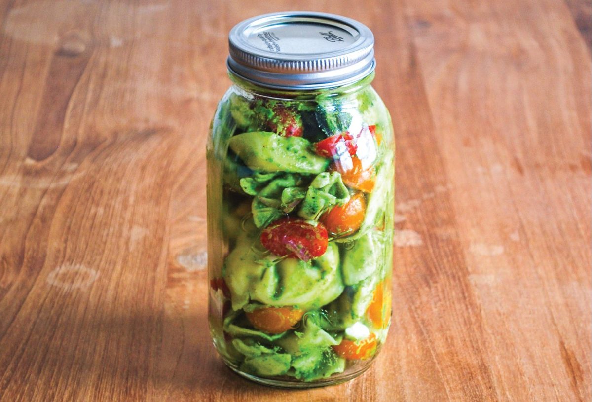 A mason jar filled with tortellini salad showcasing basil pesto, cherry tomatoes, and mozzarella.