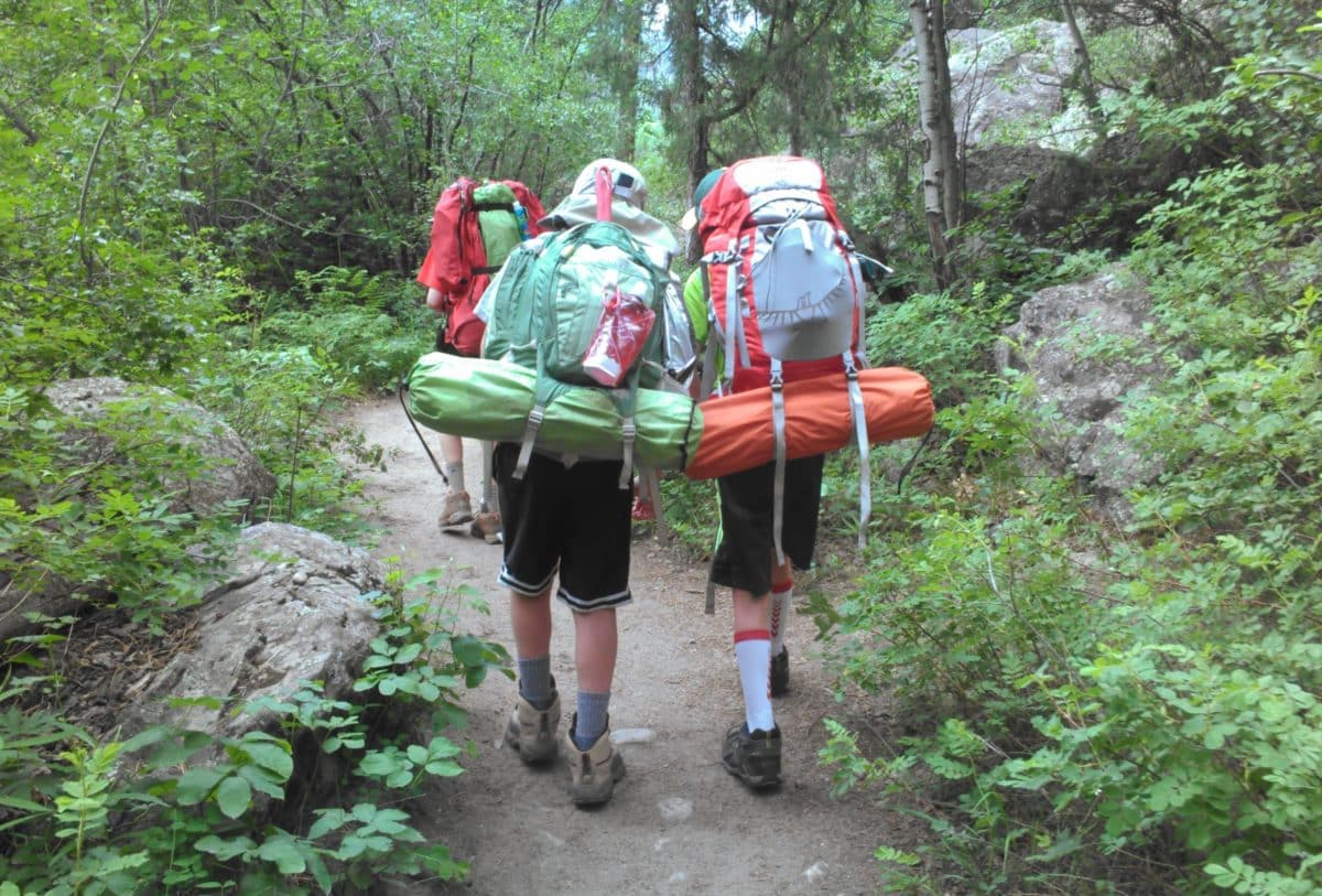 Three young boys hiking with large backpacks through a forested trail.