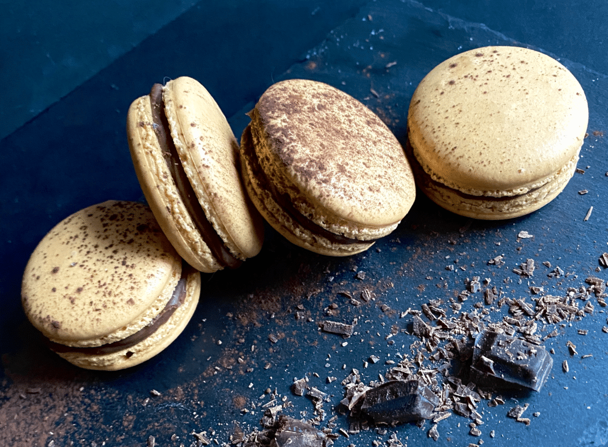 Chocolate macarons on a dark surface accompanied by dark chocolate shards.