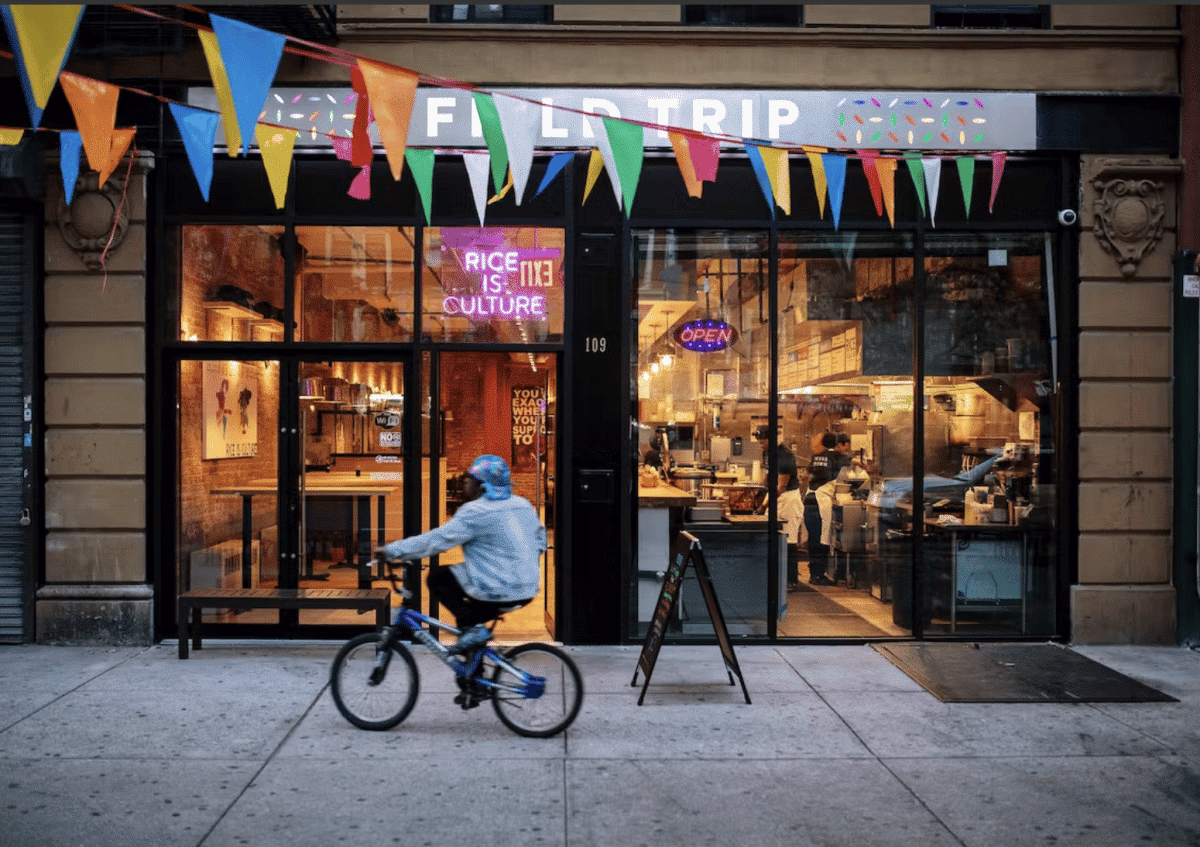 Cyclist passing by the front of FieldTripNYC with colorful banners and window displays.
