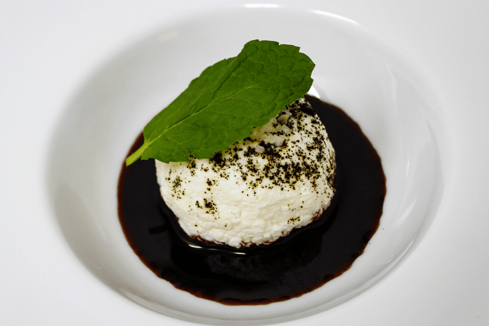 A plate with fresh cheese topped with salt ash and a port reduction, garnished with a green leaf.
